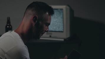 Man in White T-shirt is Sitting Against the Old Silver TV in Dark Room. Drinking Beer and Using His Smartphone. Search for TV Signal. Distortion on the Screen. Back VIew, Medium Close-Up, Film Grain video