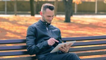 Student is Reading News or Watching on Tablet Computer in the Park in Autumn. Man in Black Leather Jacket is Sitting on a Bench Using Big White Tablet PC. Middle Close-Up Shot video