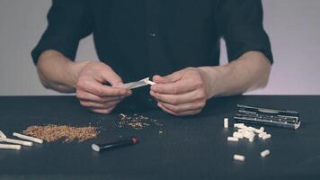 Man in Black Clothing is Rolling a Hand Made Cigarette Using Papers, Filters and Tobacco. A Way to Save on Tobacco Products. Tobacco Product Economy Concept - Slow Motion video