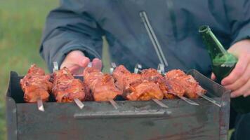 Man is Cooking Pork Meat Outdoors, Holding Beer in Green Bottle and Turning Barbecue on Skewers. Hot Grilled Kebab on the Coals. Picnic Food Preparing. Middle Close-Up Shot video