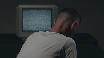 Man in White T-shirt is Sitting Against the Old Silver TV in Dark Room and Using His Smartphone. Search for TV Signal. Distortion on the Screen. Back VIew, Medium Close-Up, Static Shot, Film Grain video