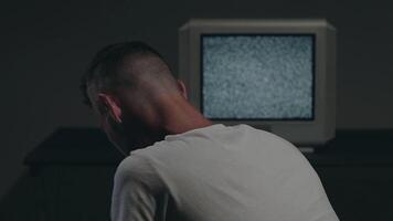 Man in White T-shirt is Sitting Against the Old Silver TV in Dark Room and Using His Smartphone. Search for TV Signal. Distortion on the Screen. Back VIew, Medium Close-Up, Static Shot, Film Grain video