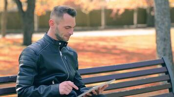 Student is Reading News or Watching on Tablet Computer in the Park in Autumn. Man in Black Leather Jacket is Sitting on a Bench Using Big White Tablet PC. Middle Close-Up Shot video