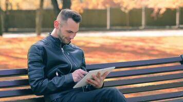 Student is Reading News or Watching on Tablet Computer in the Park in Autumn. Man in Black Leather Jacket is Sitting on a Bench Using Big White Tablet PC. Middle Close-Up Shot video