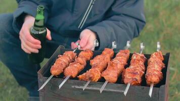 Man is Cooking Pork Meat Outdoors, Holding Beer in Green Bottle and Turning Barbecue on Skewers. Hot Grilled Kebab on the Coals. Picnic Food Preparing. Middle Close-Up Shot video