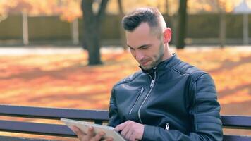 Student is Reading News or Watching on Tablet Computer in the Park in Autumn. Man in Black Leather Jacket is Sitting on a Bench Using Big White Tablet PC. Middle Close-Up Shot video