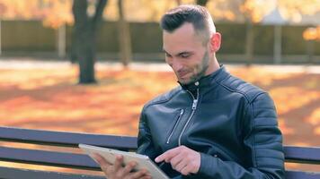 Student is Reading News or Watching on Tablet Computer in the Park in Autumn. Man in Black Leather Jacket is Sitting on a Bench Using Big White Tablet PC. Middle Close-Up Shot video