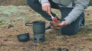 uomo nel un' foresta è cucinando pasta con in scatola stufato utilizzando un' piccolo cucinare impostare. turista nel un' escursione è preparazione per il il pranzo. avvicinamento Visualizza, statico tiro video