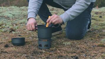 uomo nel un' foresta è cucinando pasta con in scatola stufato utilizzando un' piccolo cucinare impostare. turista nel un' escursione è preparazione per il il pranzo. avvicinamento Visualizza, statico tiro video
