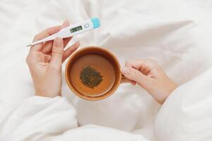 Ill patient lying in bed with a cup of tea and a thermometer on white sheets. a person feeling photo