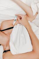 Ill woman lying in bed with a thermometer in her mouth, another one lying on the bedside table next photo