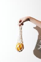 Orange in white net bag held by person in front of white wall. photo