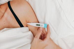 Woman lying in bed holding two thermometers, one in each hand. she appears to be checking her photo