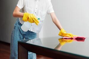 Woman cleaning glass table with yellow gloves and bottle of cleaning solution in corner of room. photo
