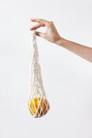 Woman's hand holding a white net bag with an orange on a white background, close up. a fresh and photo