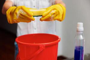 Female wearing yellow gloves holds a sponge above a red bucket and a bottle of cleaning solution. a photo