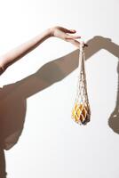Hand holding orange in front of white wall, with net bag hanging and casting a shadow. photo