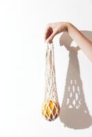 White macrame bag with orange inside held by a person, casting a shadow on a white wall. photo
