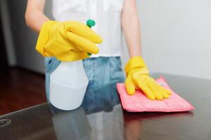 Cleaning woman holding solution bottle wearing yellow gloves on glass table with rag. photo