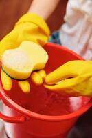 Woman in yellow rubber gloves holding a sponge in a red bucket with a yellow sponge in the center. photo