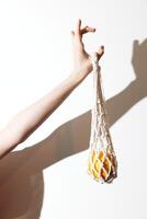 Man holding up a white bag with an orange fruit inside, casting a shadow against a white wall. photo