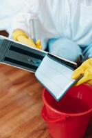 Person wearing yellow rubber gloves holding a tablet next to a bucket with red bucket on the floor. photo