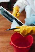 Person wearing yellow rubber gloves using a tablet on a wooden floor next to a bucket of water and photo
