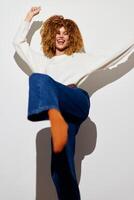 A cheerful woman with curly hair dressed in a white sweater and blue flared jeans, playfully kicking photo