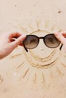 Hands holding sunglasses with smiley face drawn in sand in front of person wearing sunglasses on the photo