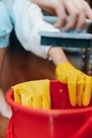 Cleaning person wearing yellow rubber gloves scrubbing a red bucket with a brush on wooden floor photo
