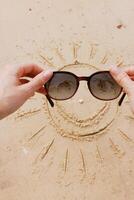 Hands holding sunglasses on a sandy beach with a drawn sun and smiley face. beach accessories photo