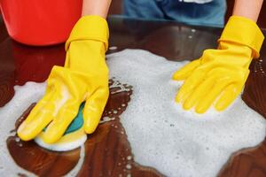 Woman cleaning table with soap and sponge on wooden surface, wearing yellow gloves. bucket of water photo