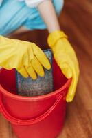 Person in rubber gloves holding bucket with sponge on hardwood floor in front of another person in photo