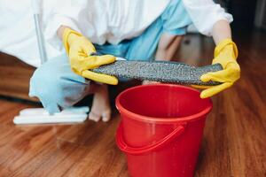 Person wearing rubber gloves using mop and bucket to clean floor in front of red pail. photo