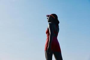 A stylish woman in a red swimsuit stands confidently against a bright blue sky, promoting a sense of photo