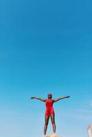 A confident woman stands with arms outstretched on a rock, wearing a red swimsuit, with an expansive photo