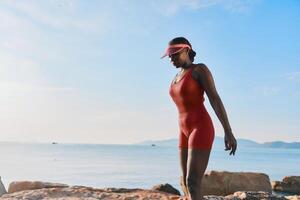 A confident woman in a red swimsuit, wearing a visor, enjoys a peaceful stroll along the beach, with photo