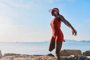 A fit woman performs a yoga pose on rocky terrain by the sea, dressed in a vibrant red outfit with a photo