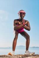 A female athlete in a pink outfit poses confidently on the sand with a serene ocean backdrop photo