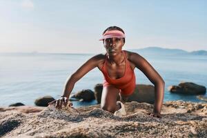 A fit woman in a red outfit and visor performs a push up on rocky terrain by the water, with a calm photo