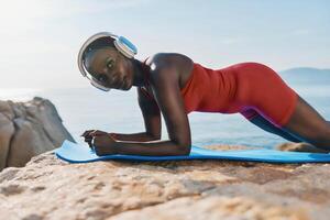 Fit woman doing a plank on a blue mat, surrounded by serene seaside scenery and soft waves in the photo