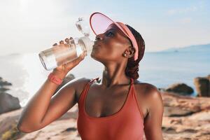 Athletic woman refreshing herself with water by the beach, wearing a cap and tank top, enjoying a photo