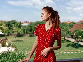 A woman in a red polka dot dress enjoys a peaceful garden view from a sunny balcony. photo