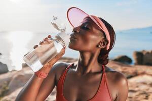 A fit woman in a cap enjoying water from a bottle, with a serene ocean background and a warm, sunny photo
