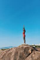 A confident woman poses atop a large rock, raising her arms in celebration or joy, with a bright photo