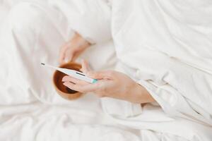 Man using thermometer to measure temperature of coffee cup on bedside table with white sheet. photo
