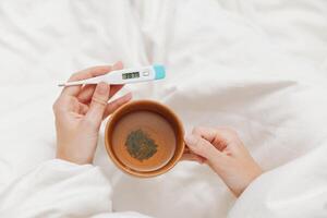 Sick person lying in bed holding a thermometer and a cup of tea, with another thermometer on the photo