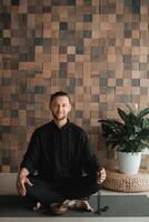 Portrait of a young man in a black kimano sitting in a lotus position on a gym mat in the interior photo