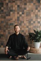 Portrait of a young man in a black kimano sitting in a lotus position on a gym mat in the interior photo