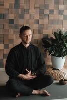 Portrait of a young man in a black kimano sitting in a lotus position on a gym mat in the interior photo